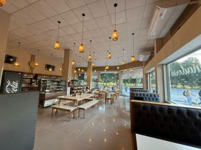Interior de una cafetería con mesas, sillas y lámparas colgantes, junto a un gran ventanal.