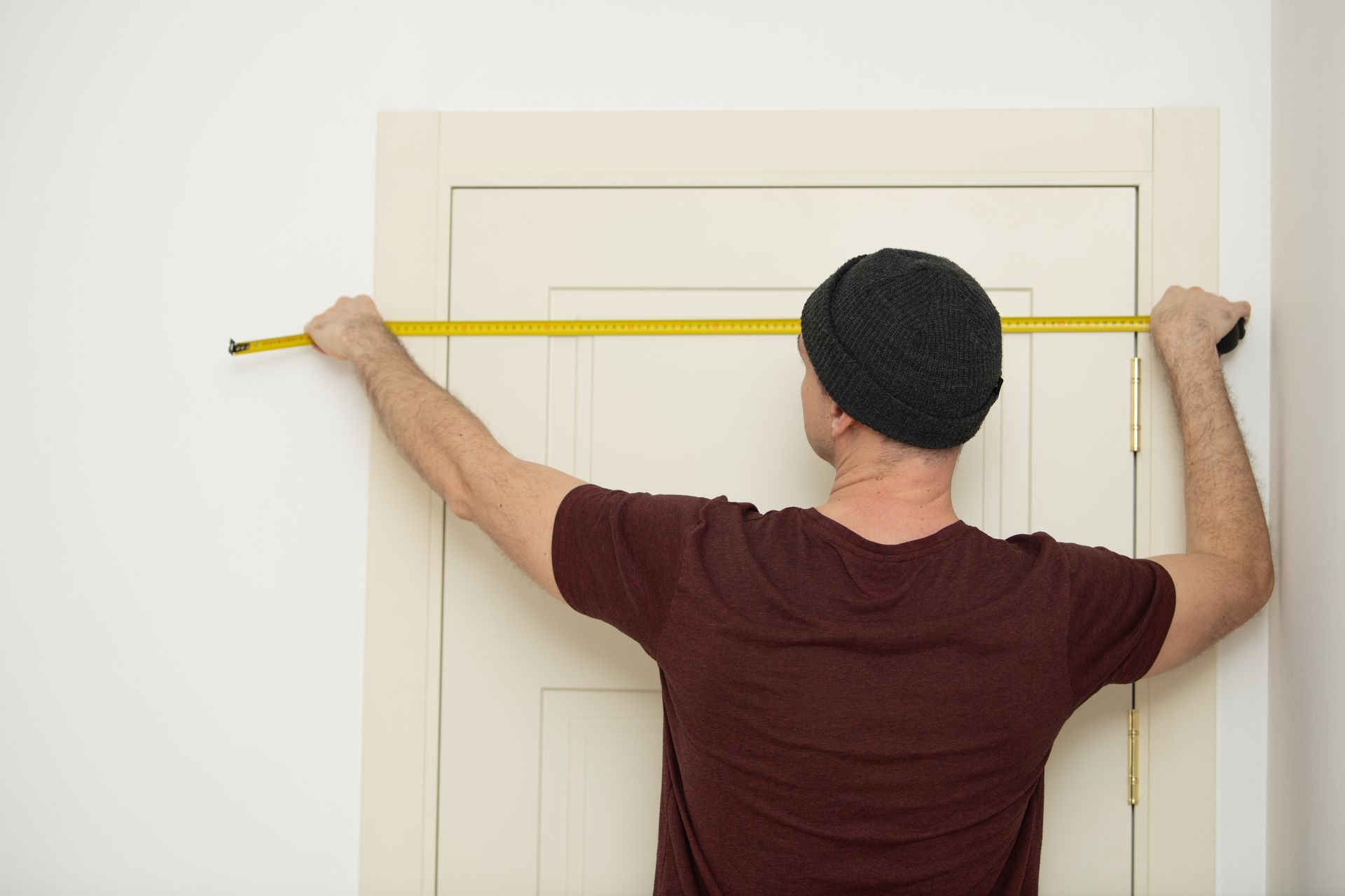 Man wearing a beanie measuring the width of a beige door with a yellow tape measure.