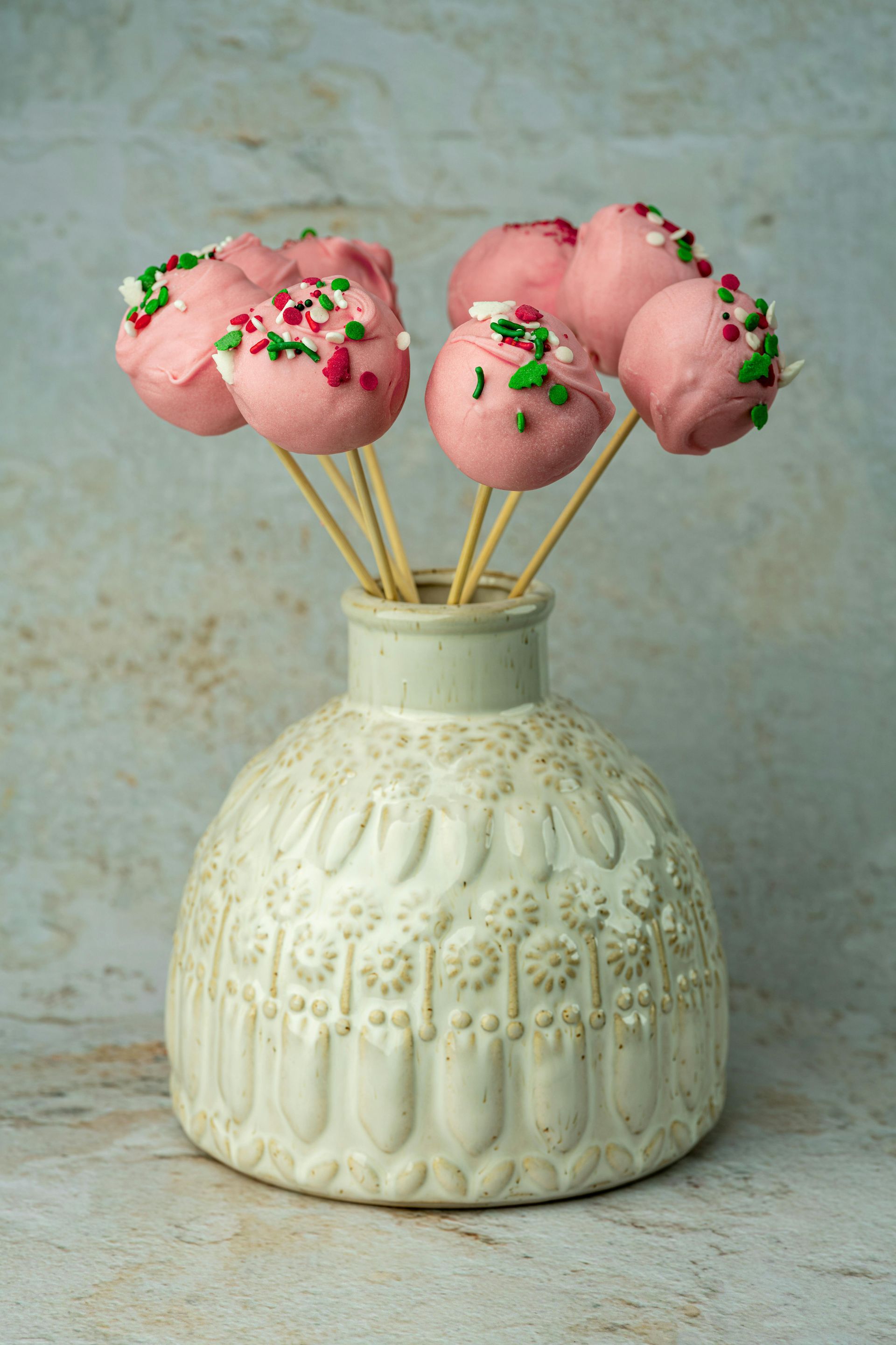 Roze cakepops op stokjes in een decoratieve witte vaas, versierd met kleurrijke hagelslag.