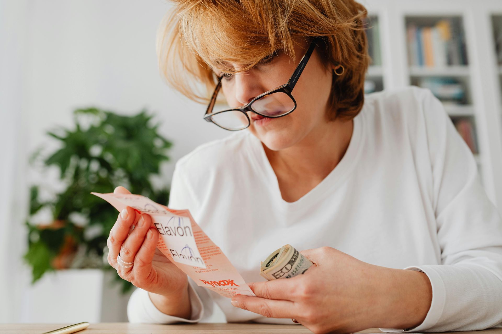 Woman with glasses examines a lottery ticket, holding money in the other hand.
