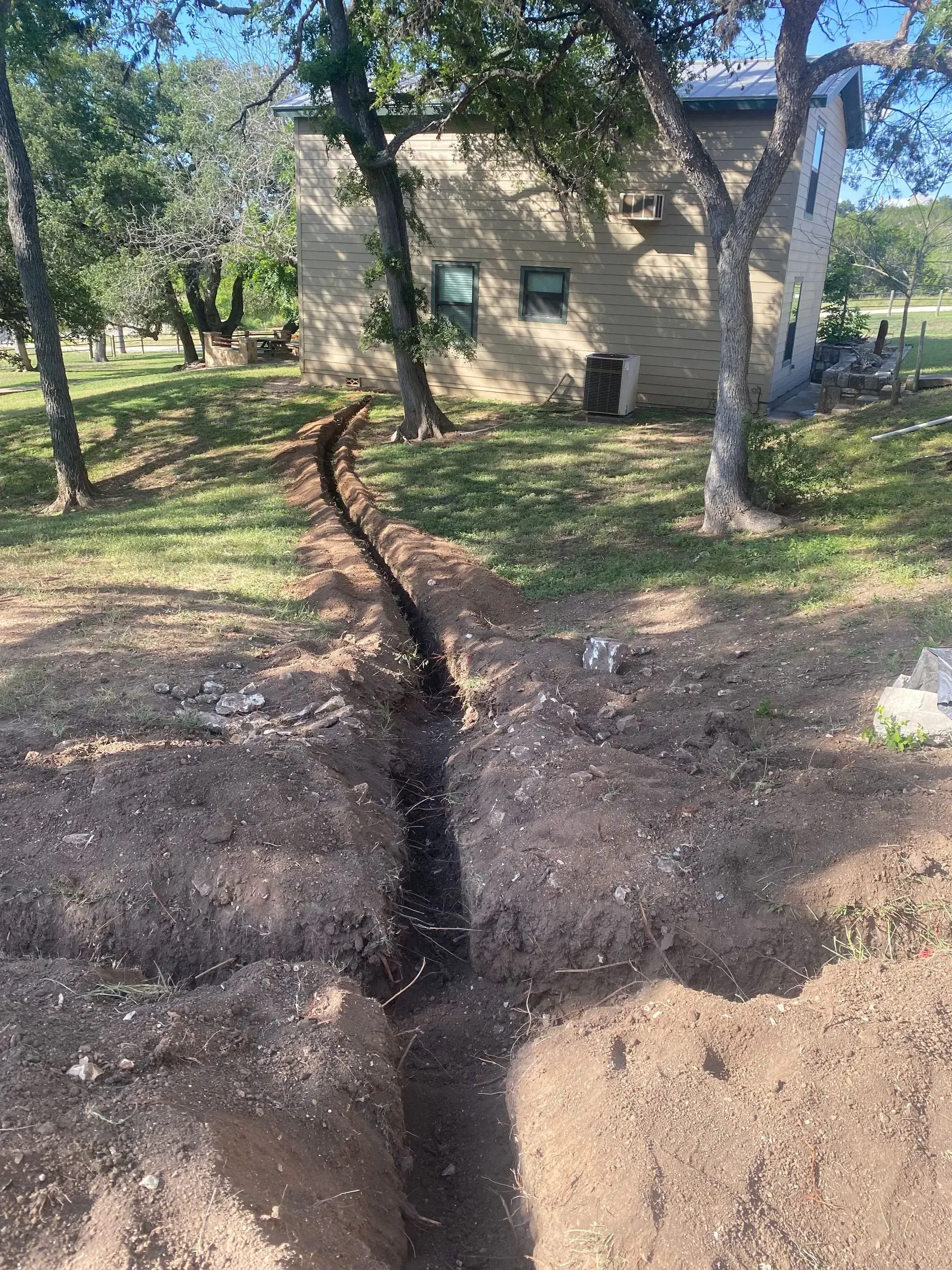 A freshly dug trench in a grassy yard, leading towards a stone building under a sunny sky.