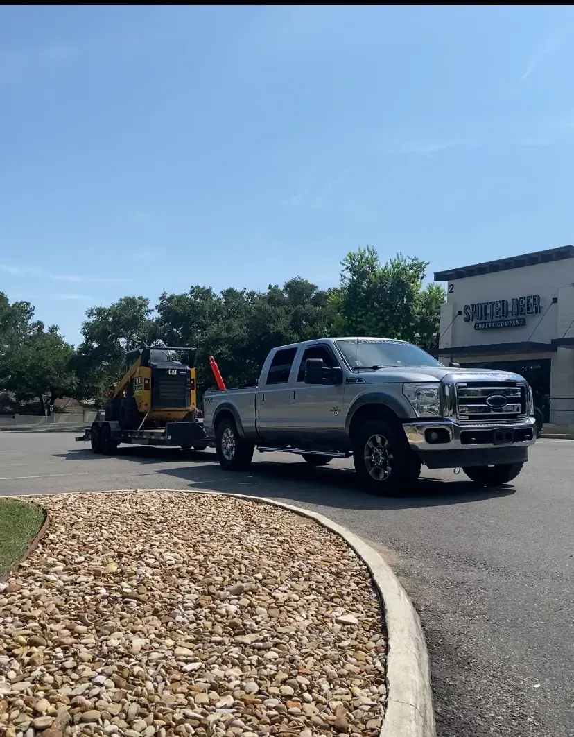 Silver pickup truck towing construction equipment on a trailer in front of a building.