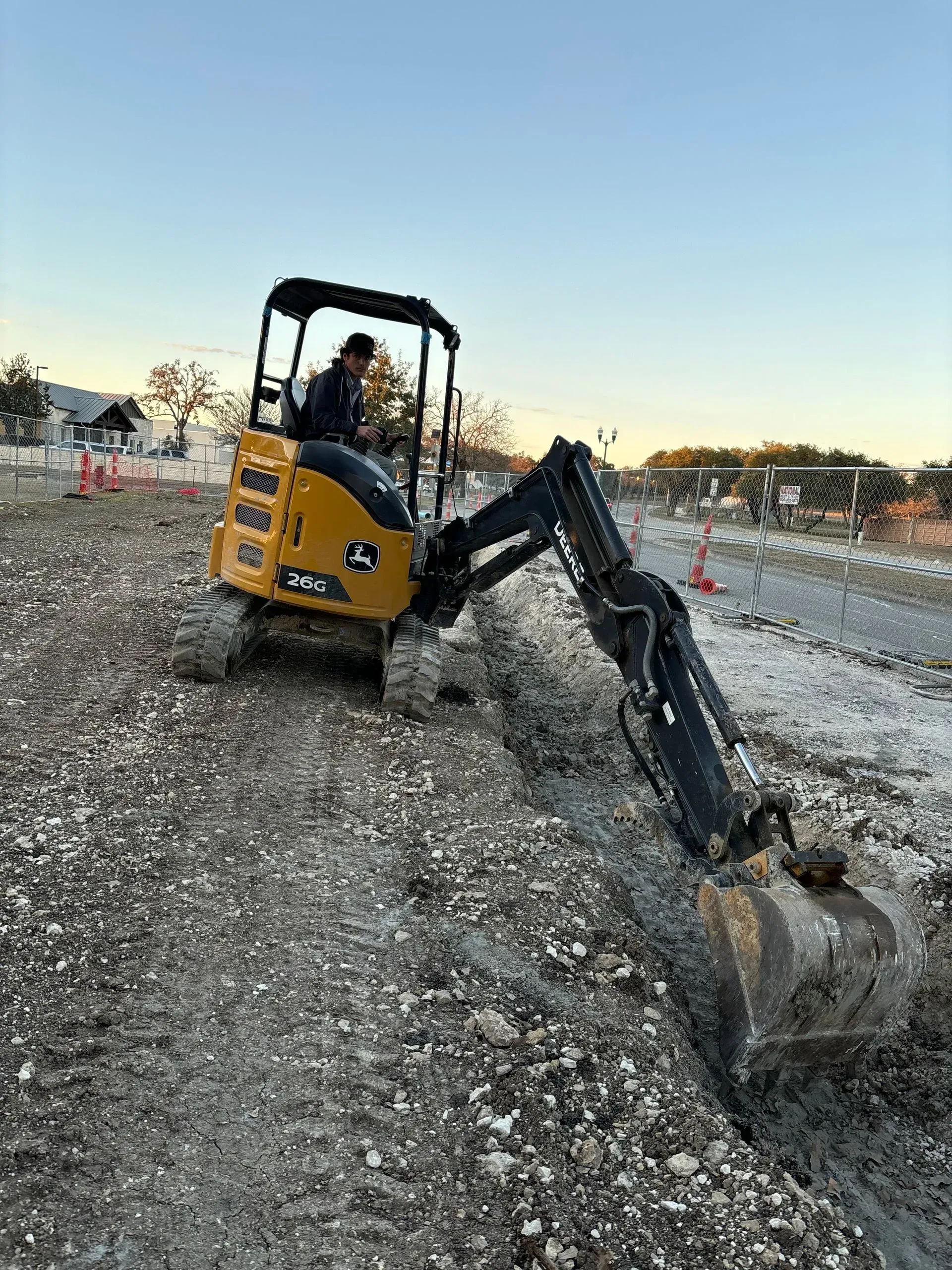 A person operates a yellow John Deere mini-excavator digging in a construction zone.