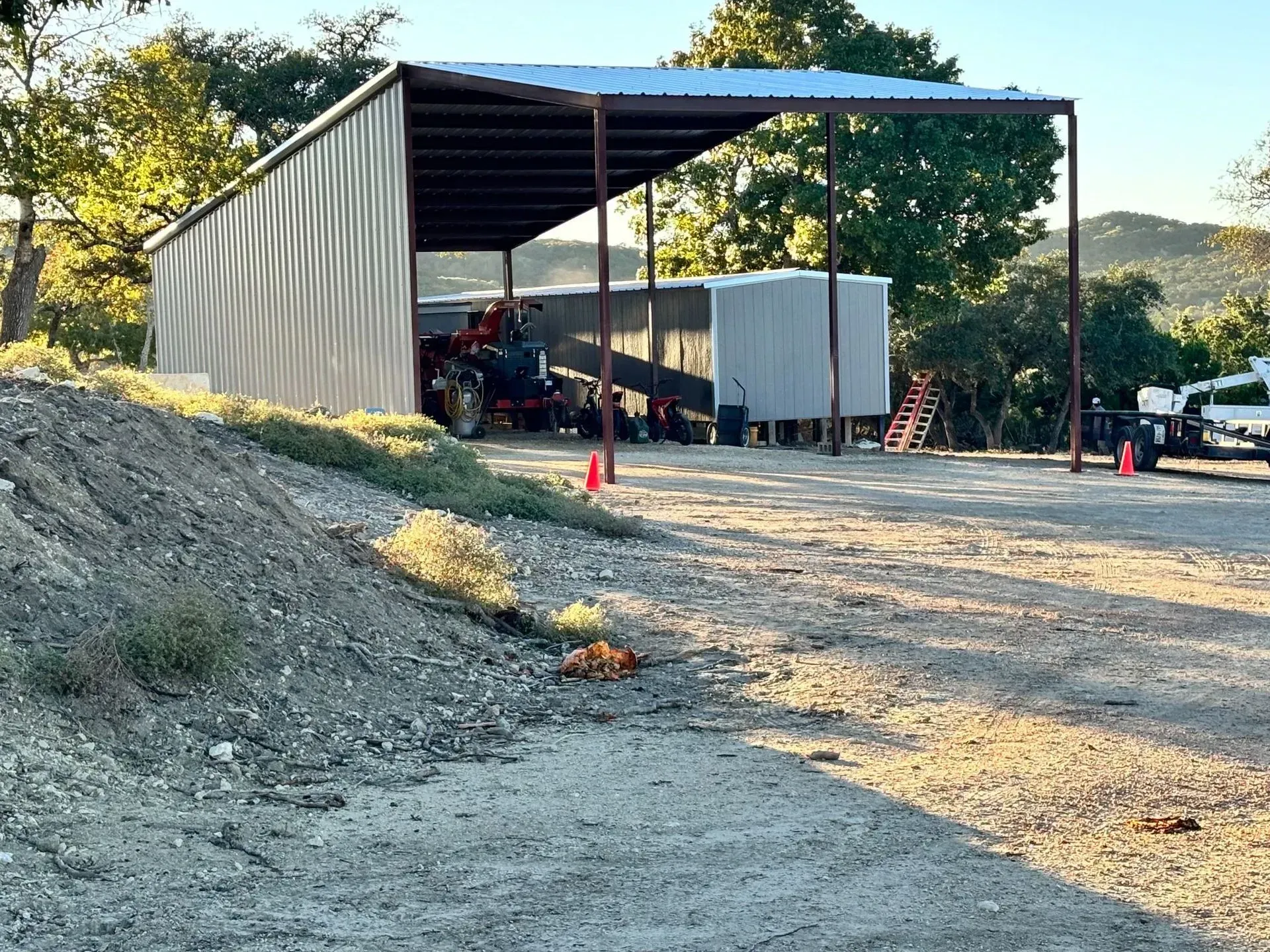 Metal shed with a tractor parked inside, on a gravel lot near trees.