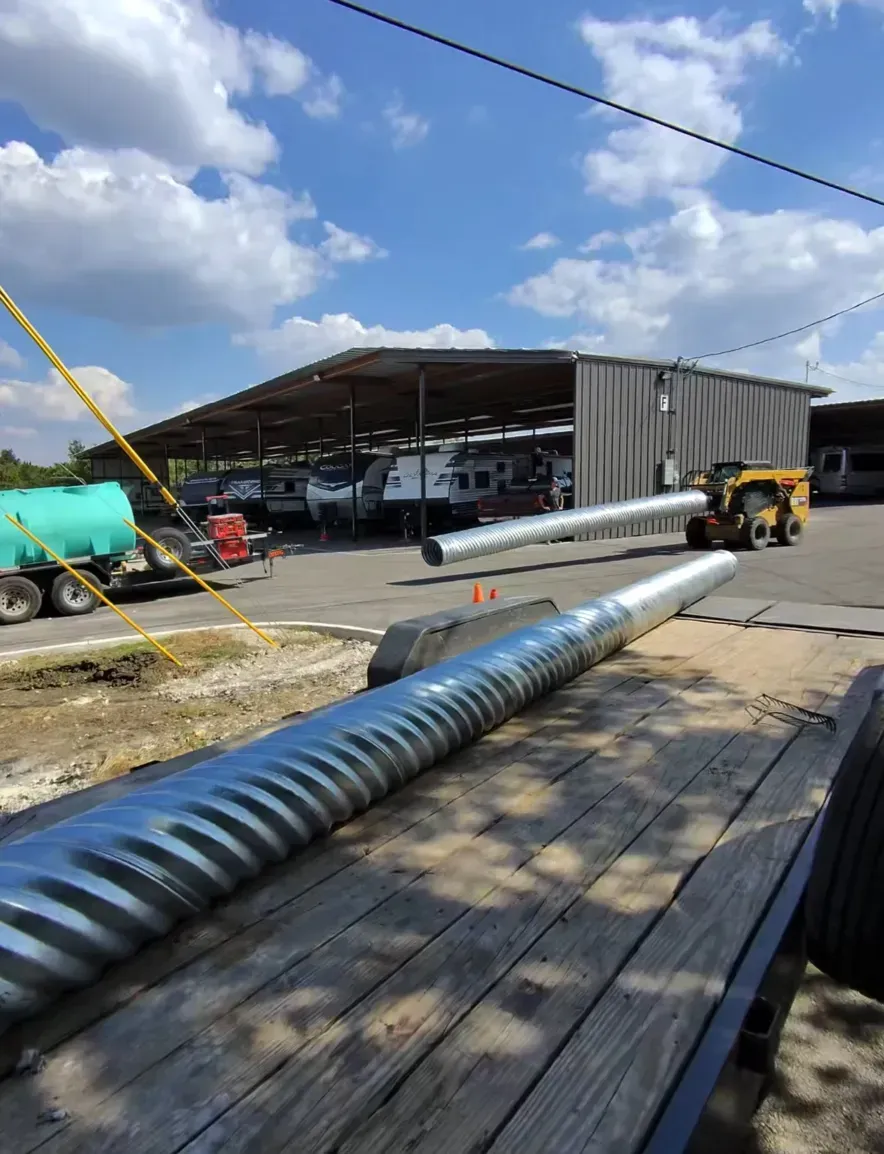 A metal culvert pipe being lifted by a small yellow tractor, with another on a trailer. 