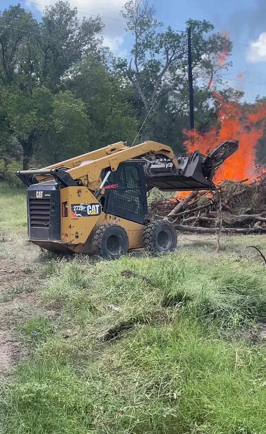 Yellow skid steer loader by a brush fire, working in a grassy field.