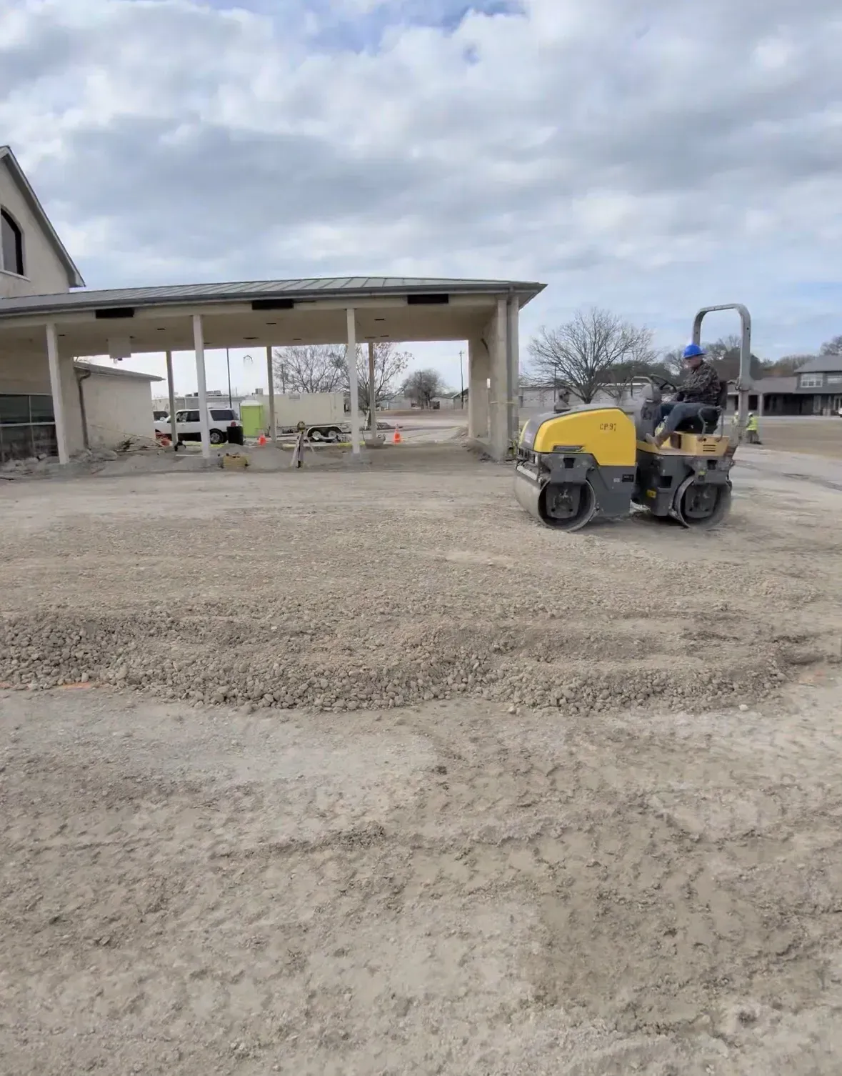 A person operating a road roller on a gravel surface near a building under a cloudy sky.