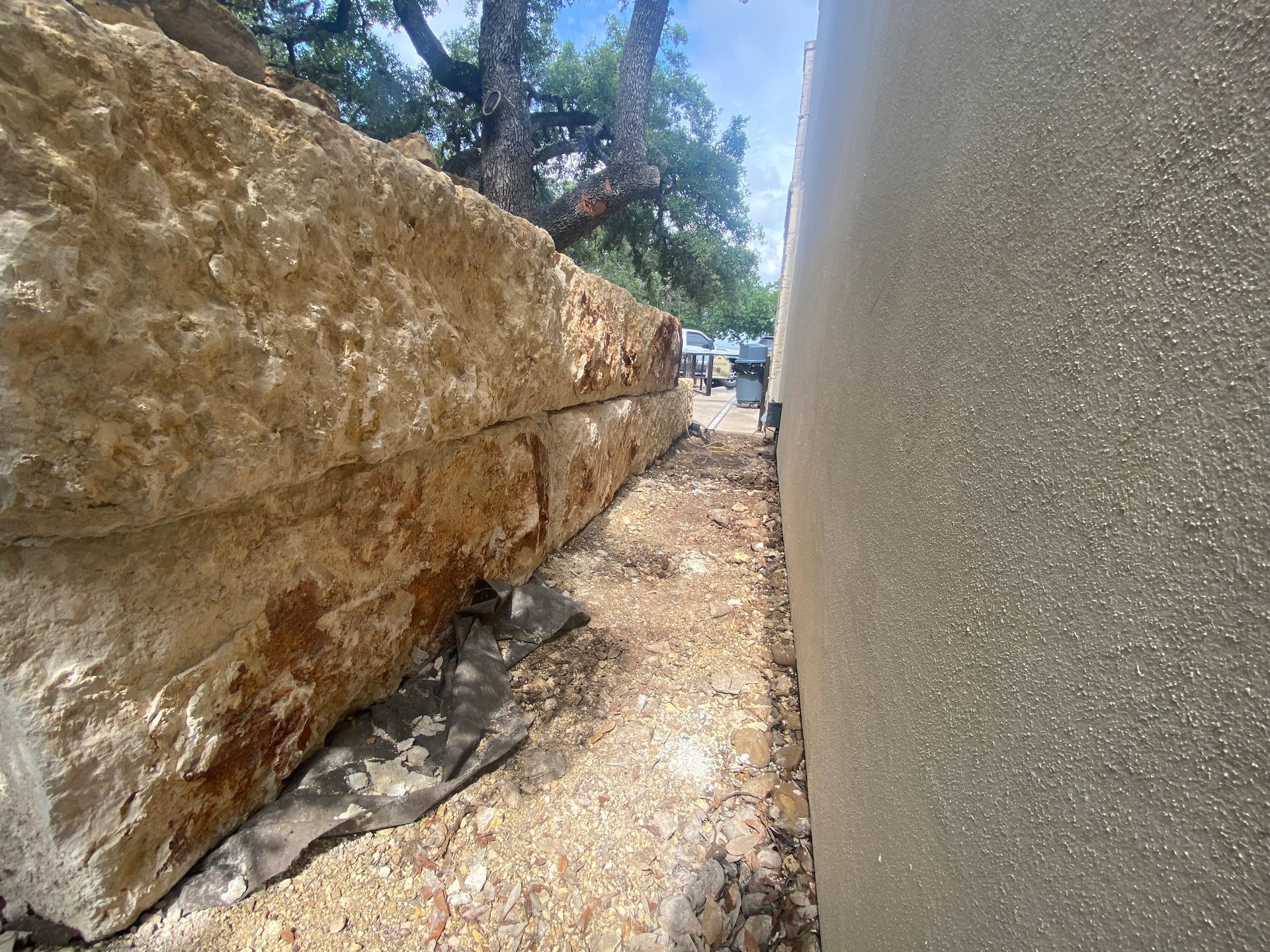 Large stone wall next to a stucco wall, with a narrow walkway in between.