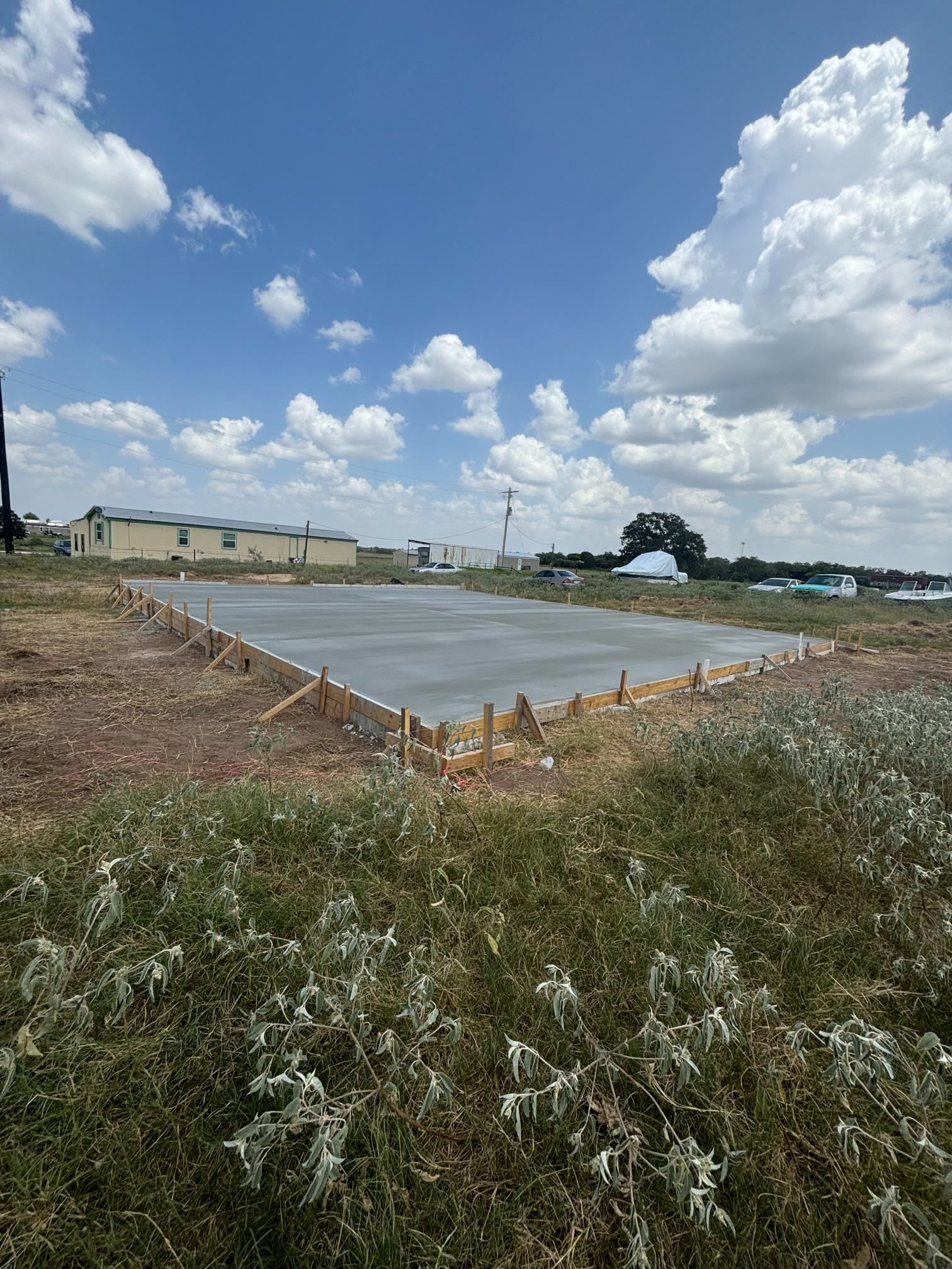 Concrete slab foundation under construction in a grassy field beneath a bright blue sky with clouds