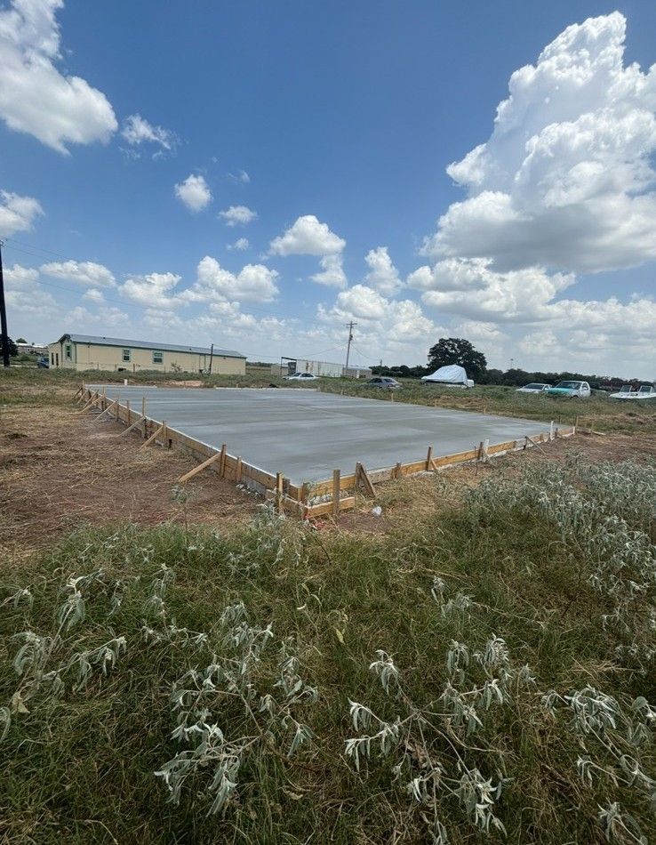 Concrete foundation slab at a construction site under a partly cloudy sky, with grass and buildings nearby.