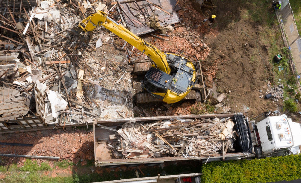Aerial view of a yellow excavator demolishing rubble beside a truck and brick wall