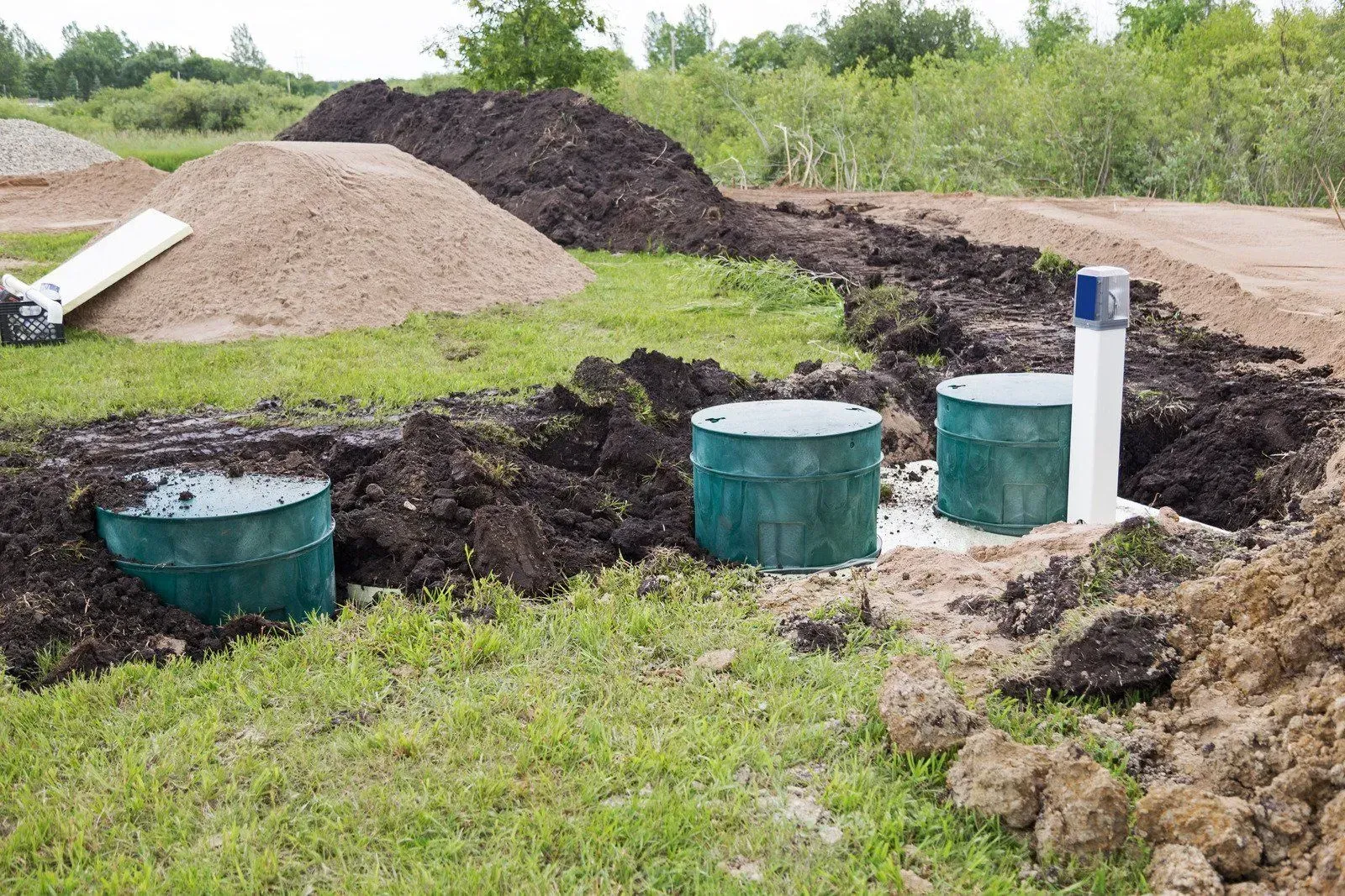 Installation of a septic system: green tanks, white pipe, dirt piles in a grassy field.