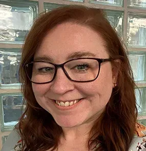 Woman with auburn hair wearing glasses smiles in front of a glass block wall.