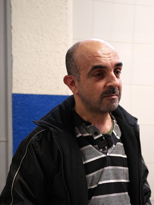 Man in a dark jacket and striped shirt standing indoors against a blue and white wall, looking to the side.