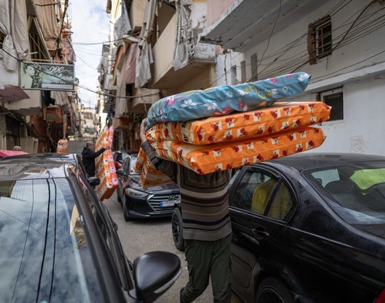 Man carrying stacked mattresses down a narrow street lined with parked cars and buildings