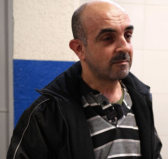 Man standing indoors against a blue-and-white wall, wearing a black jacket and striped shirt.