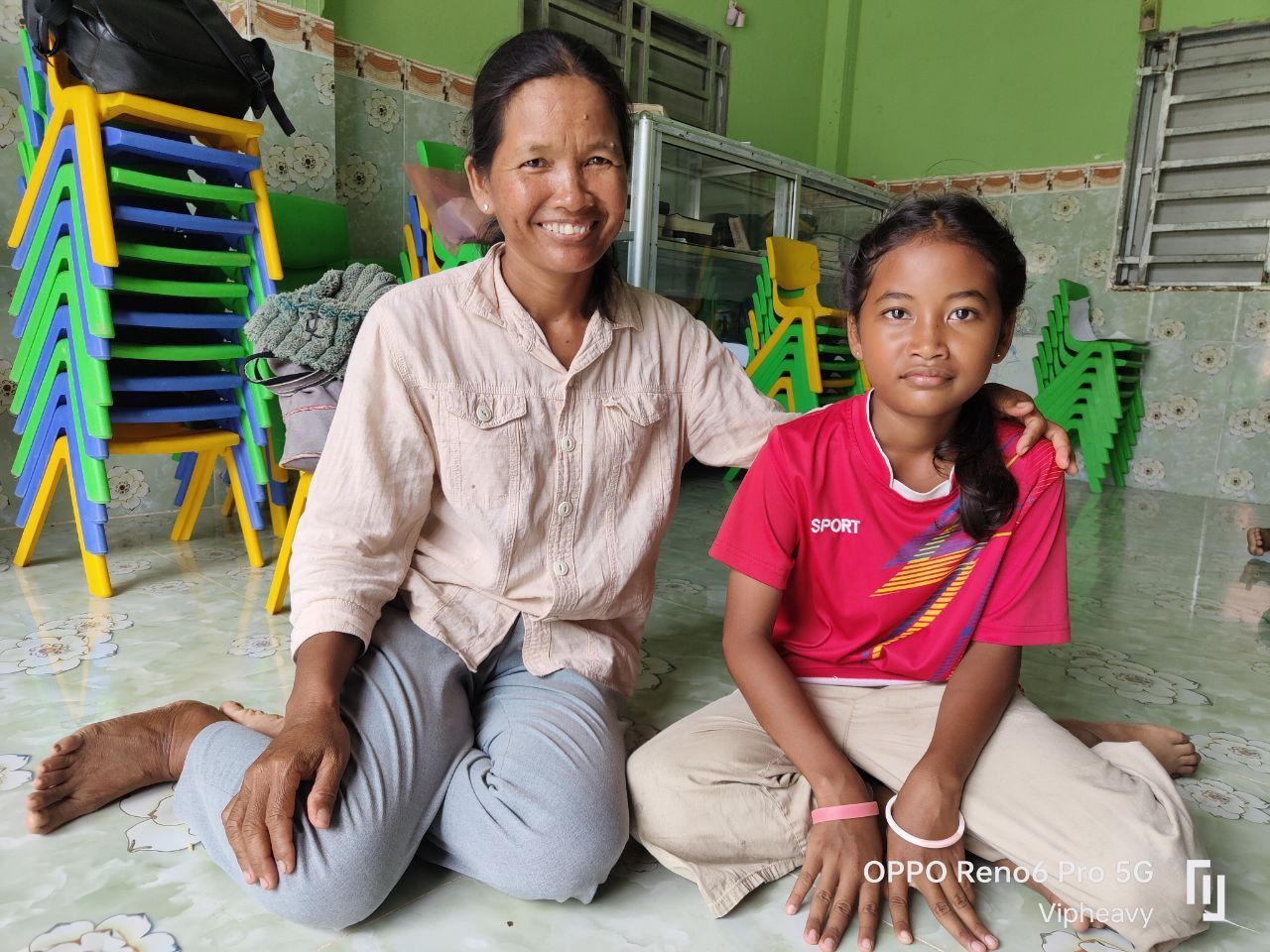 Woman with arm around a girl, both kneeling on the floor. Stacked chairs in background.
