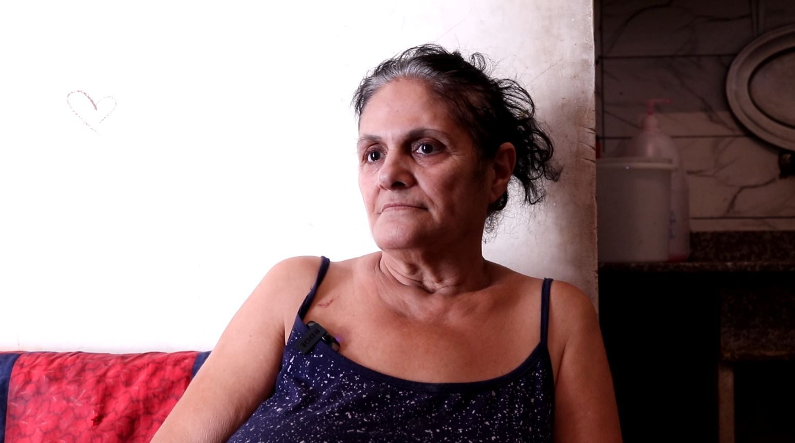 Woman in a blue top sitting in front of a white wall, looking at the camera.