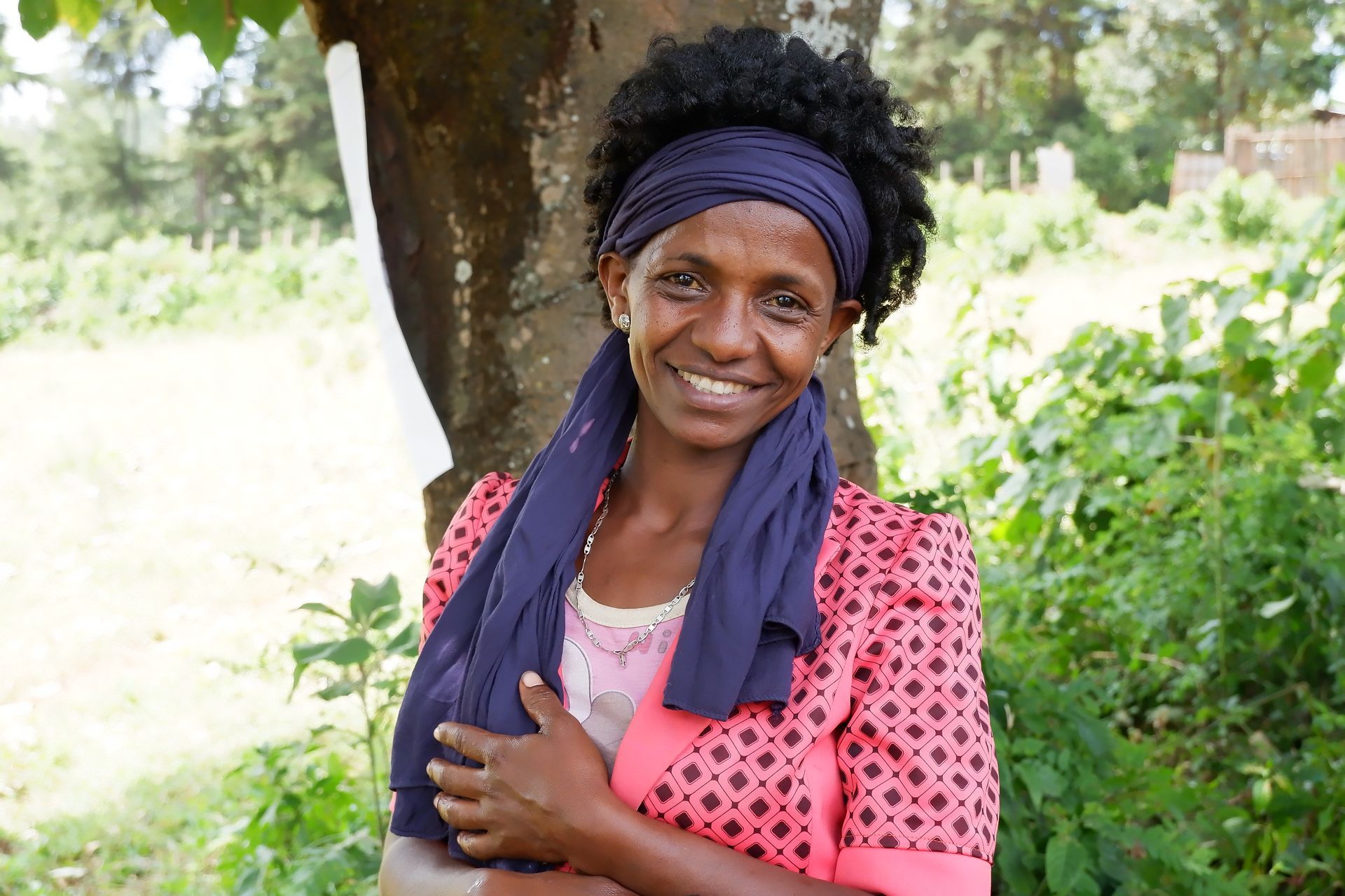 Woman smiles, wearing a pink patterned shirt, blue scarf and headband, standing outside near a tree.