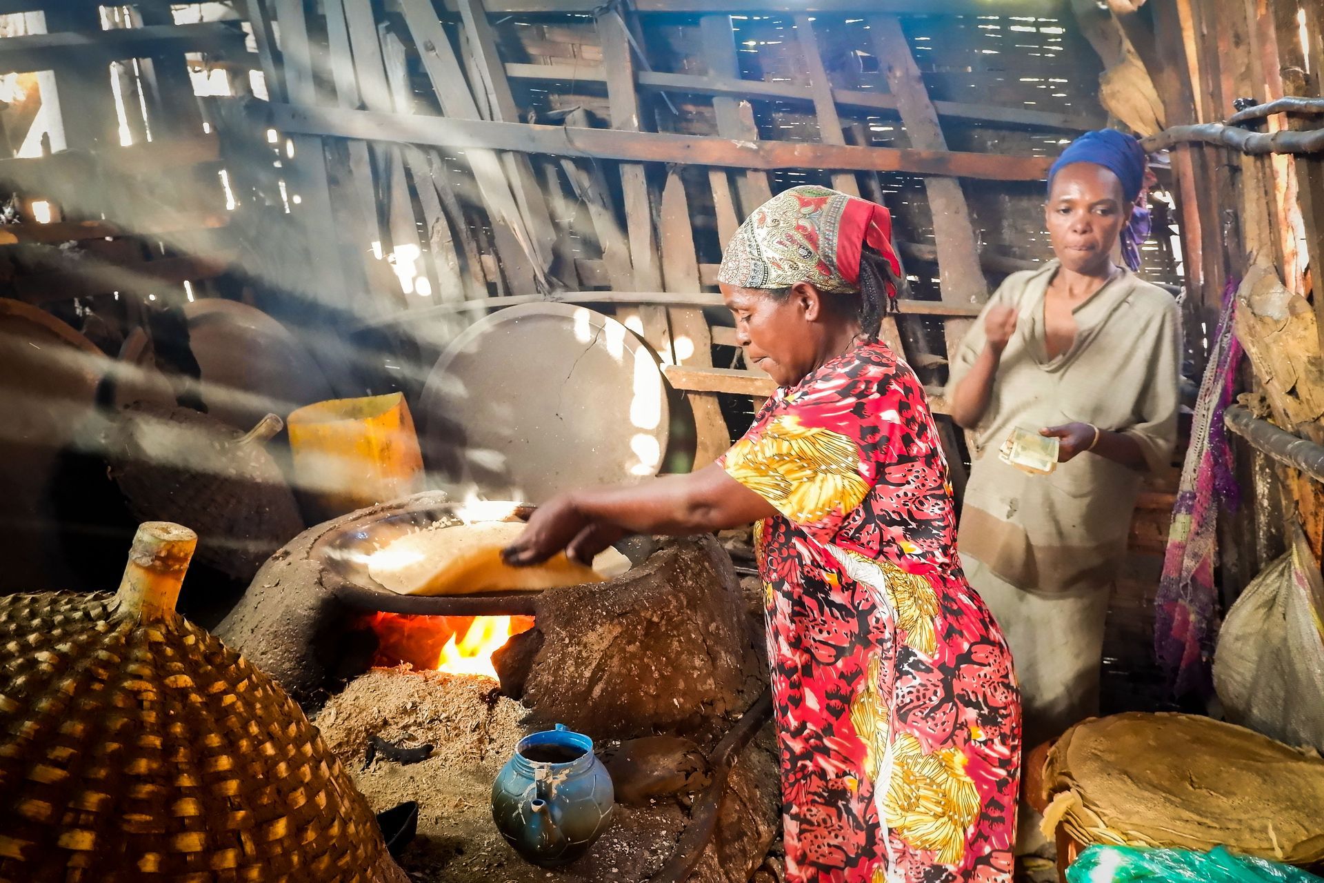 Woman placing flatbread in oven, another woman watches. Warm light fills rustic kitchen.