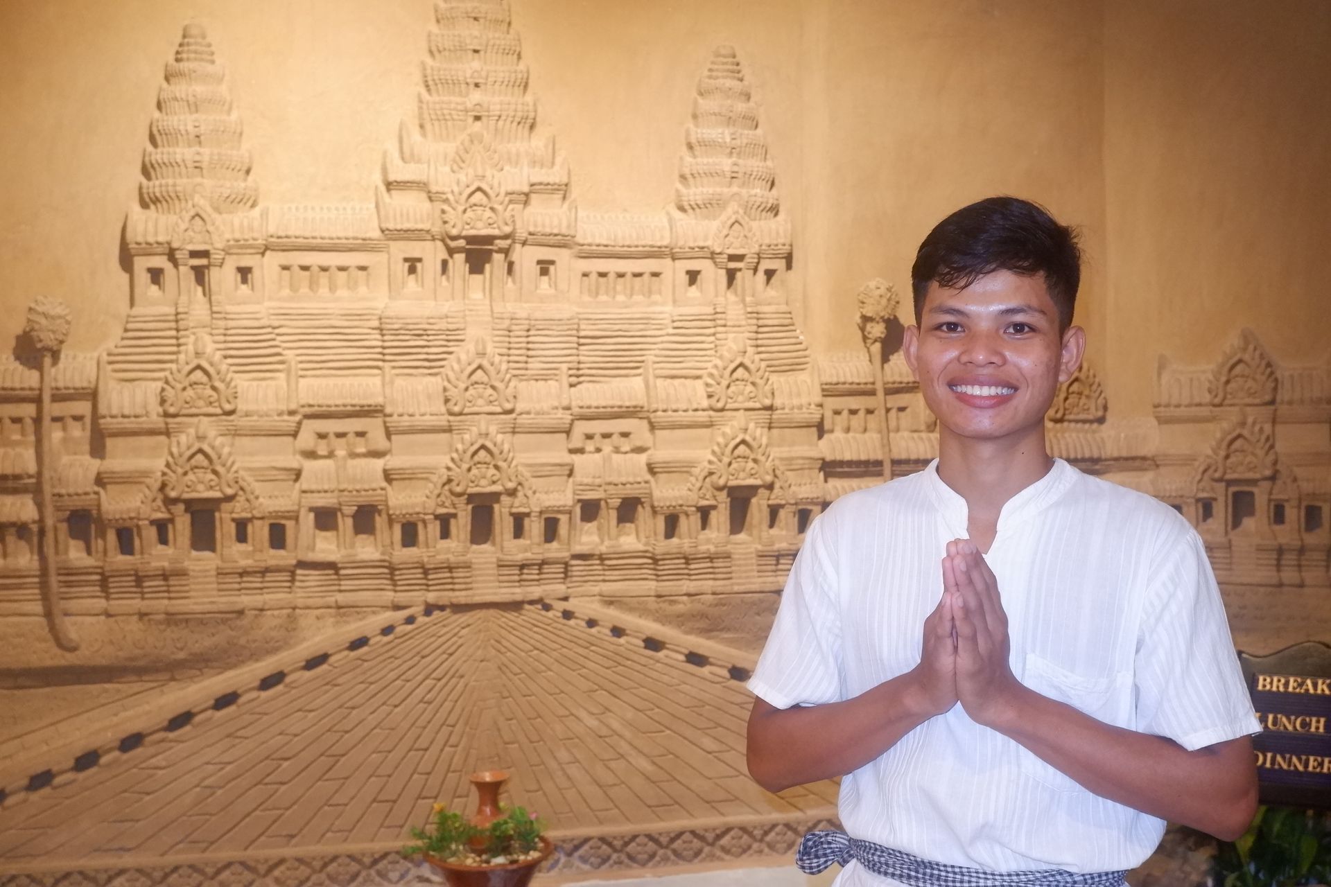 Man in white shirt, hands together in front of him, smiles in front of Angkor Wat relief.