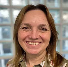 Woman smiling, wearing a floral shirt, in front of a window with white blocks.