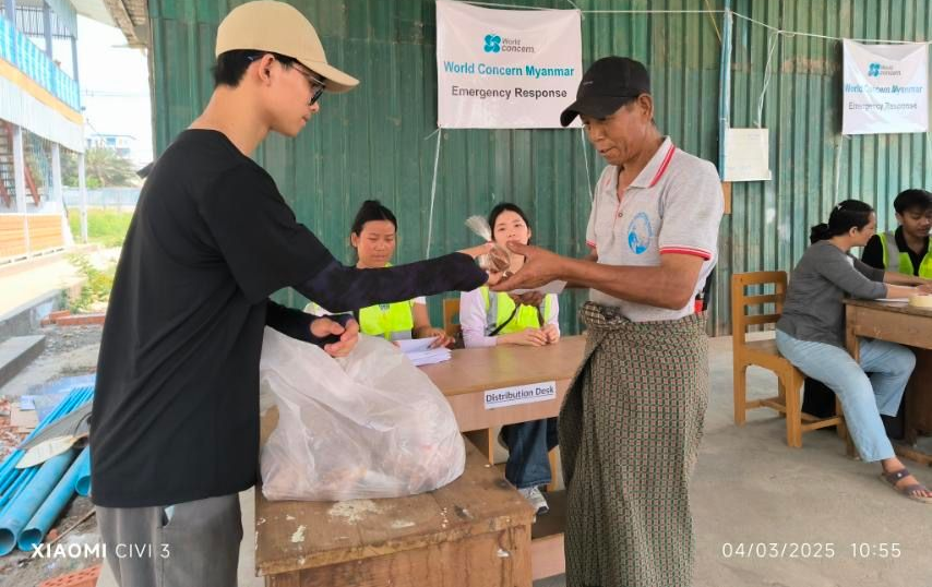 A person handing a bag of items to another person at an aid station; Red Cross signage visible.