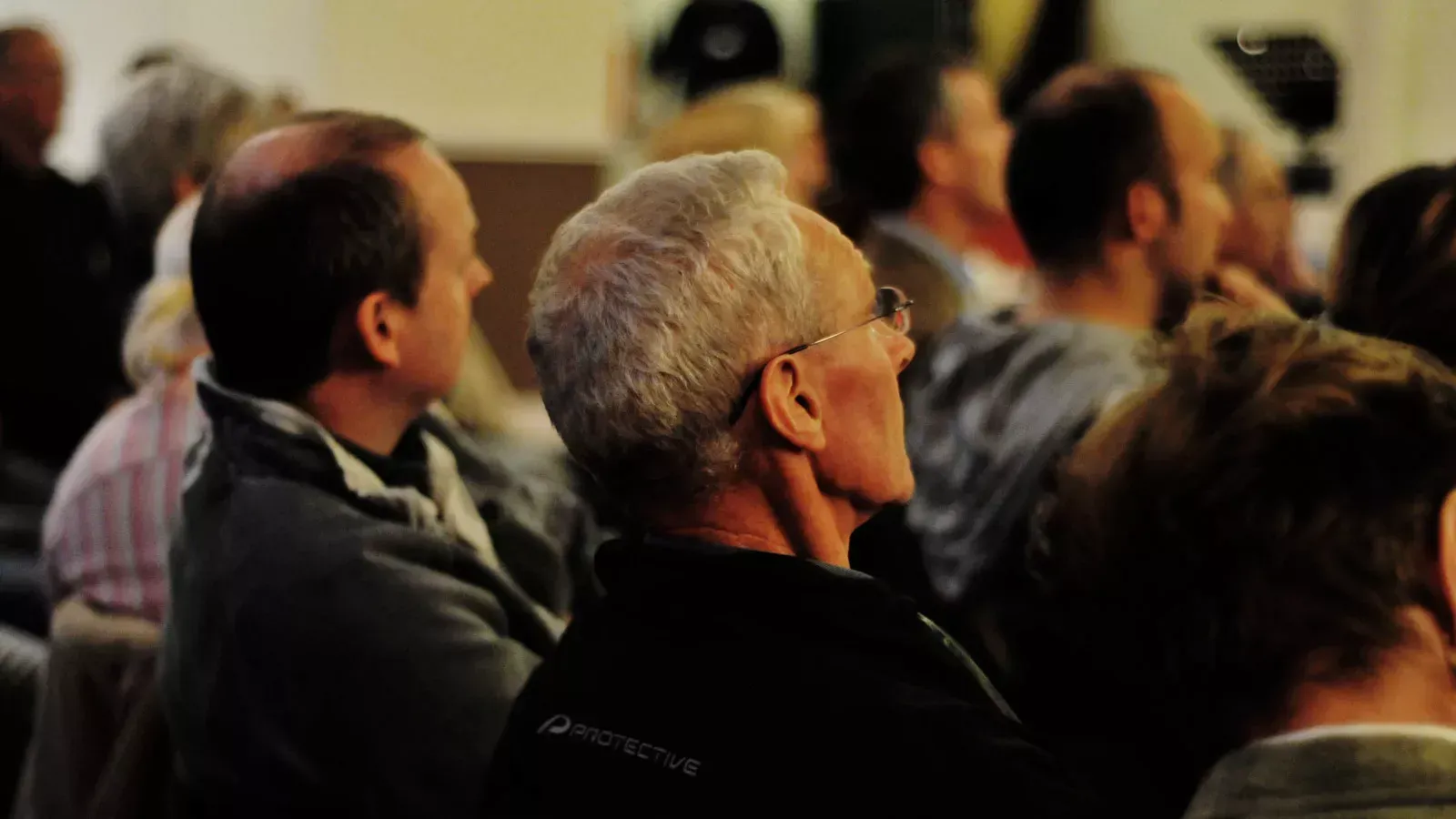 Audience at event, heads facing forward; gray-haired man in glasses in foreground, others blurred in background.
