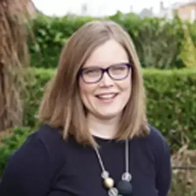 Woman with glasses smiles, wearing a dark top and beaded necklace, outdoors.
