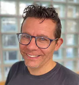 Man with glasses smiles in front of a window, wearing a dark shirt.