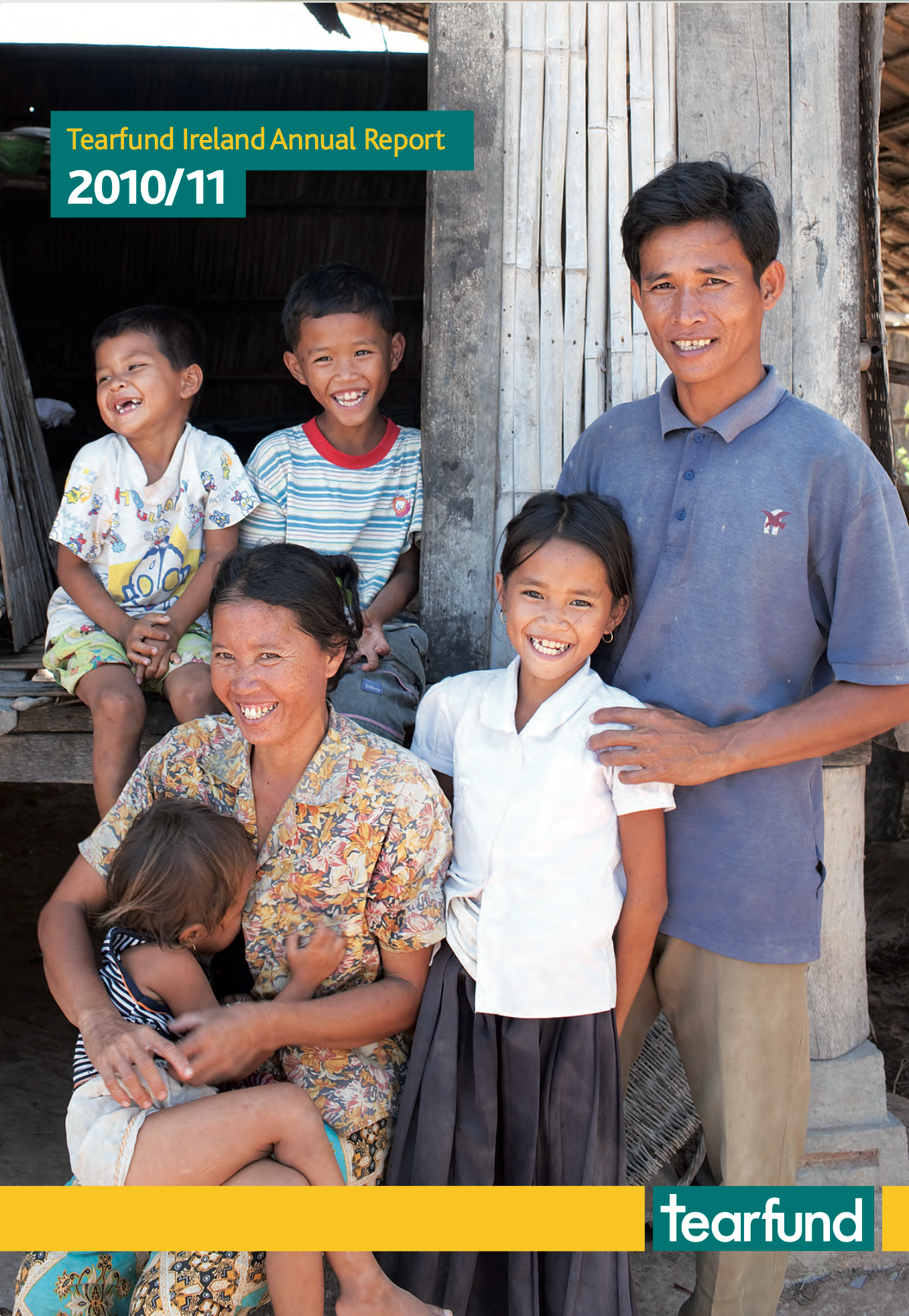 Family smiles outside a weathered building. The mother holds a child, children smile. Tearfund logo.