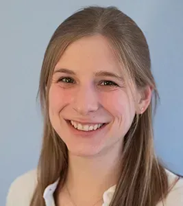 Woman with long blonde hair smiles at the camera, wearing a white shirt; light blue background.