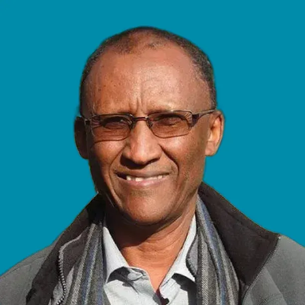 Man with glasses smiling, wearing a jacket and scarf, against a teal background.
