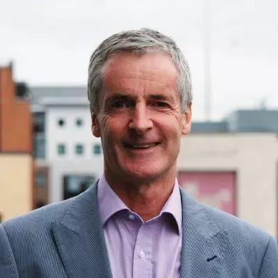 Man in a grey suit and pink shirt smiles against an urban background.