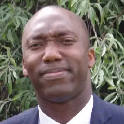 Man in suit, looking slightly to the side with a neutral expression, outdoors with leafy background.