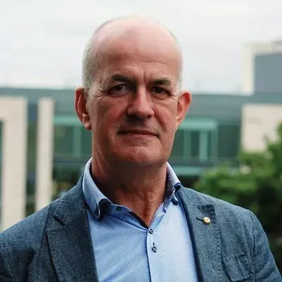 Man in a blue shirt and blazer looking at the camera outdoors, with a building in the background.