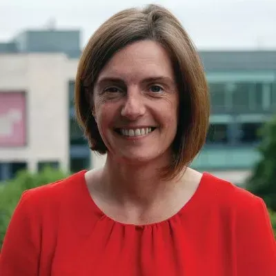 Woman with short brown hair, smiling, wearing a red top, outdoors near a building.