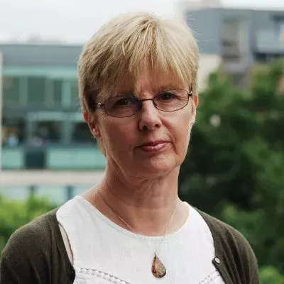 Woman with glasses, short blonde hair, wearing a white shirt and olive cardigan, outdoors.