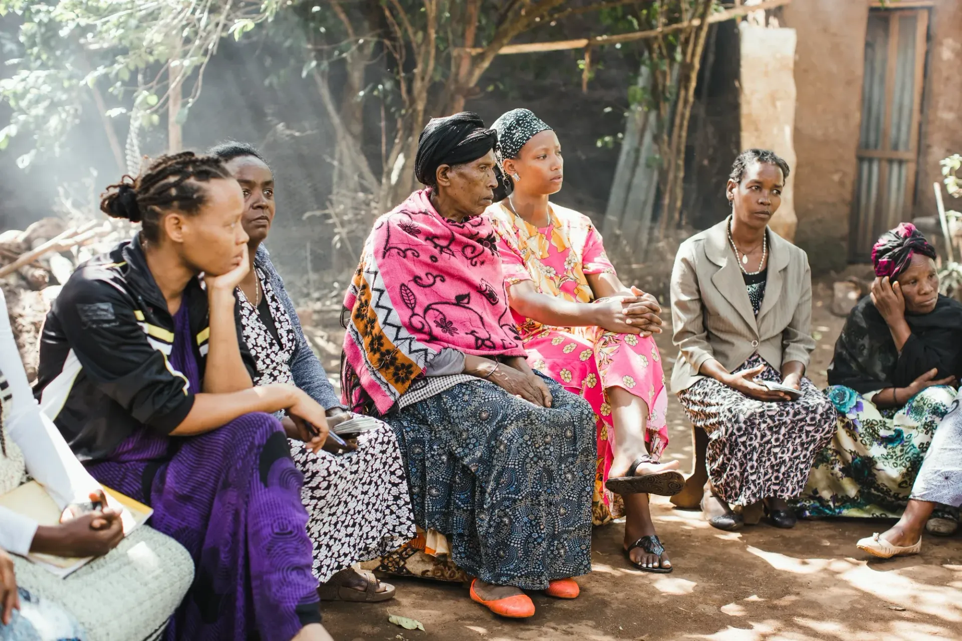 Group of people sitting outdoors, listening attentively. Some wear colorful patterned dresses.