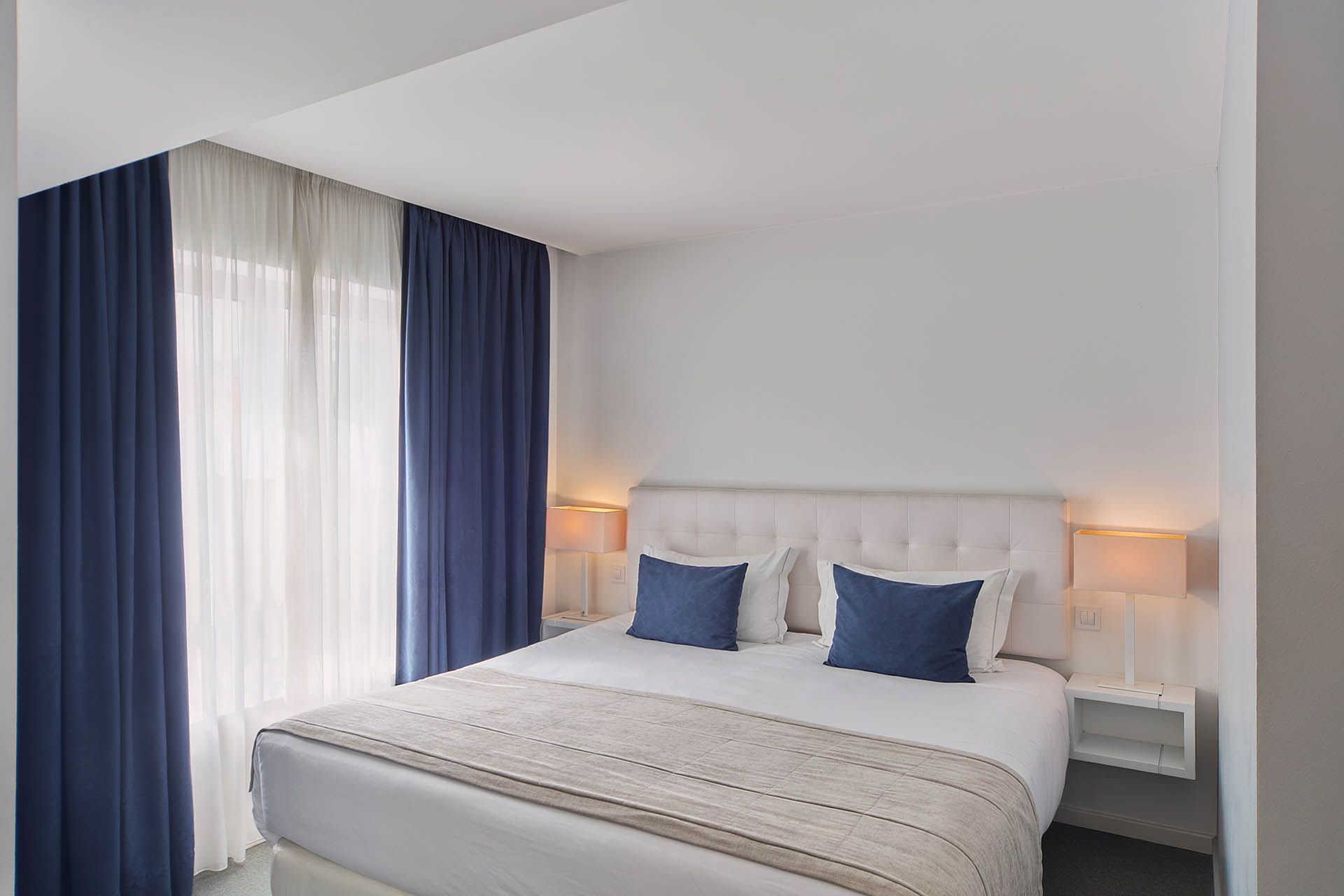 Quarto luminoso com cama branca, cortinas azuis e candeeiros de cabeceira.