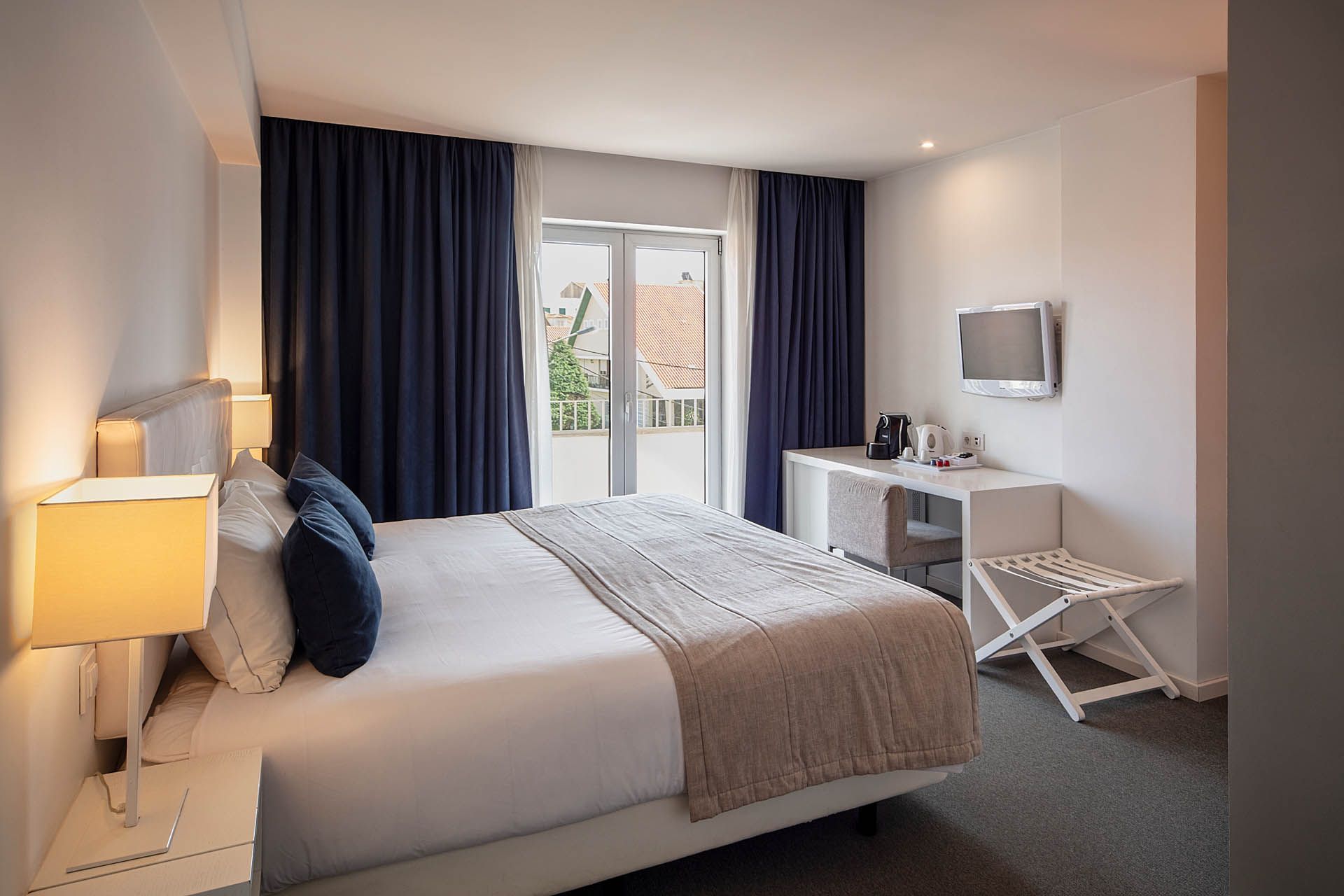 Quarto moderno e luminoso com uma cama grande, cortinas azuis, escrivaninha, cadeira, TV de parede e portas para varanda.