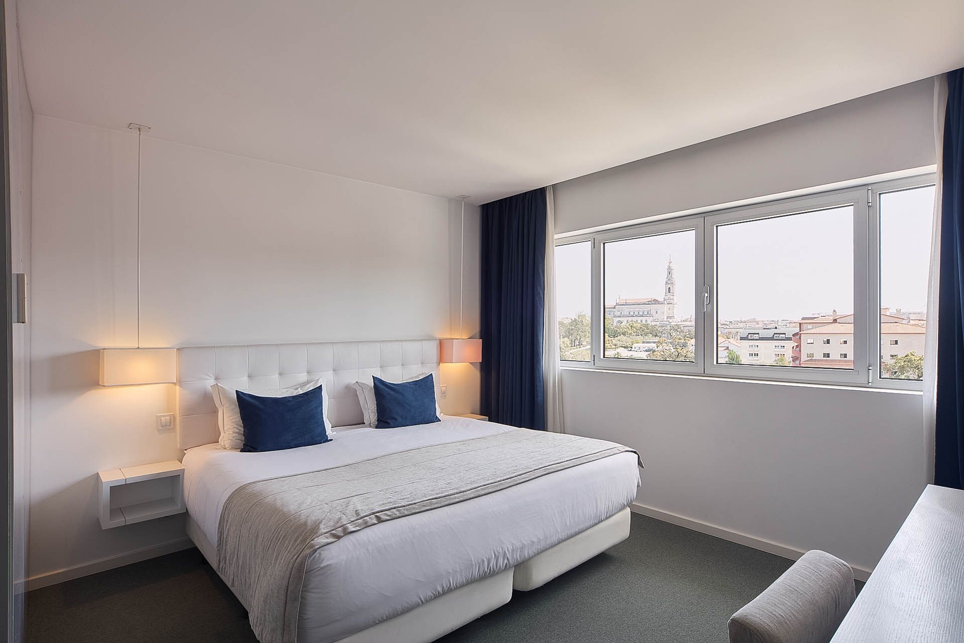 Quarto moderno com cama, candeeiros de cabeceira e uma grande janela com vista para a cidade.