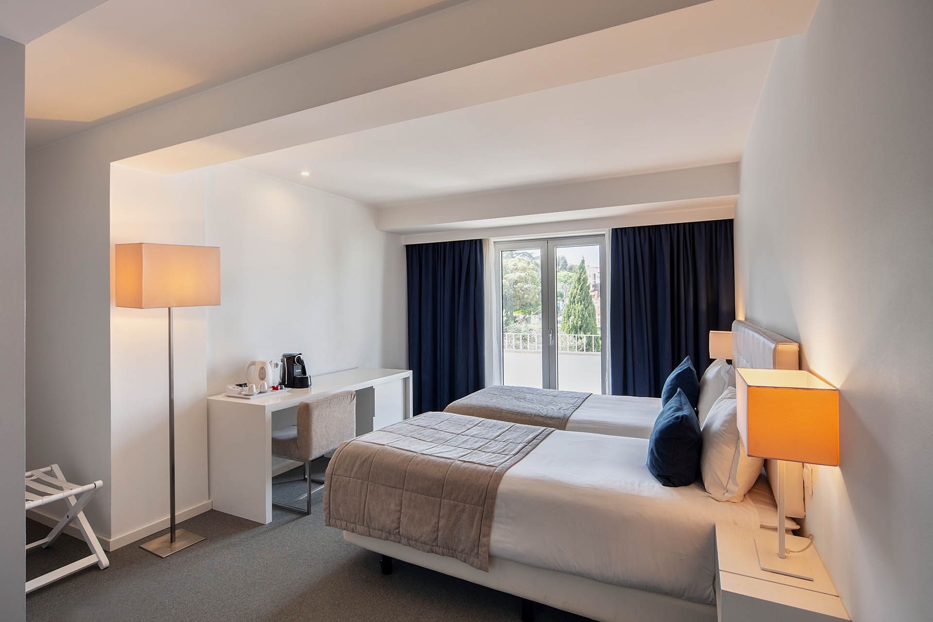 Quarto moderno com cama, abajur, escrivaninha e cortinas escuras junto à janela.