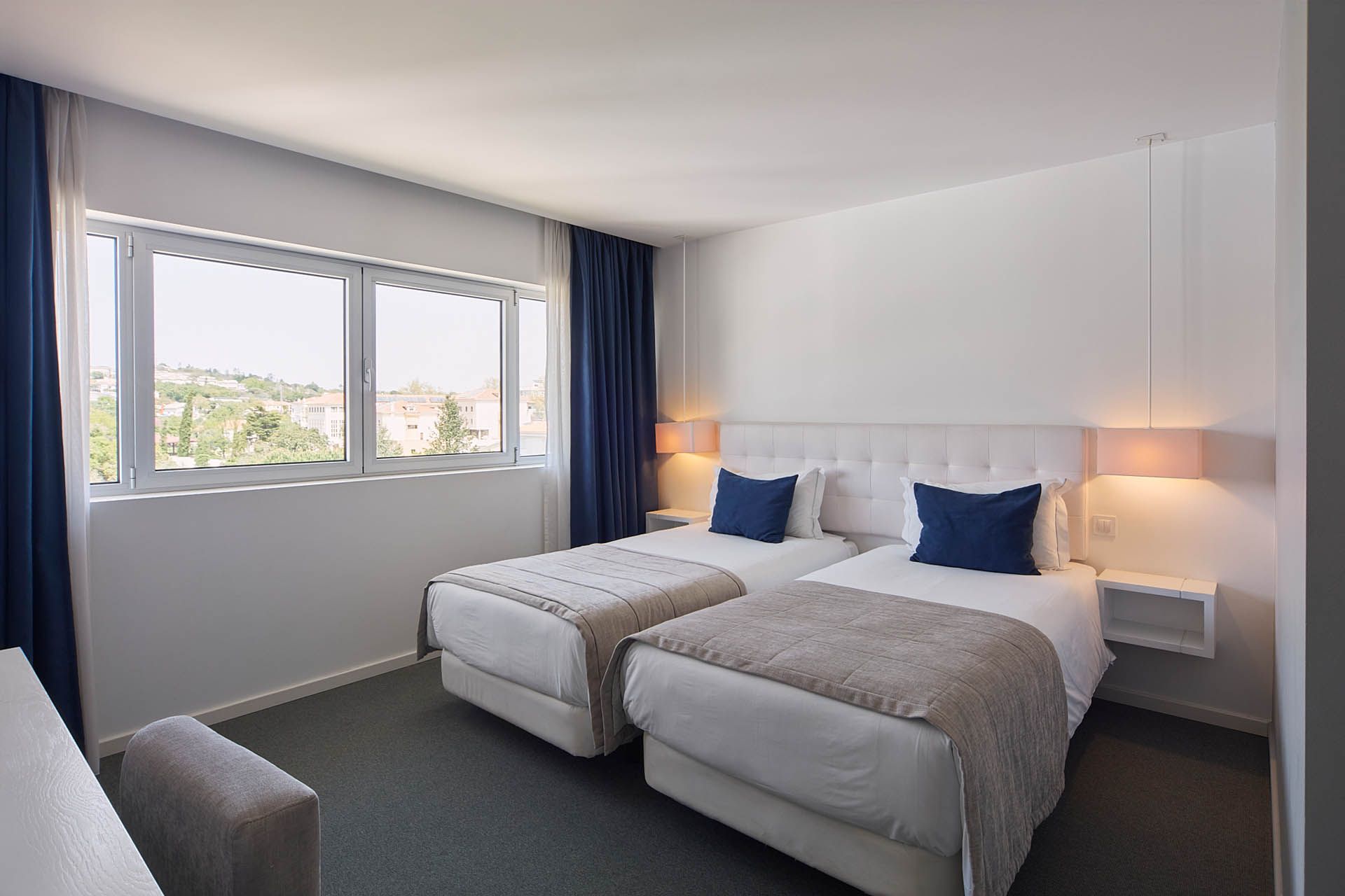 Quarto de hotel moderno com duas camas de solteiro, travesseiros azuis, abajures e janela com cortinas.