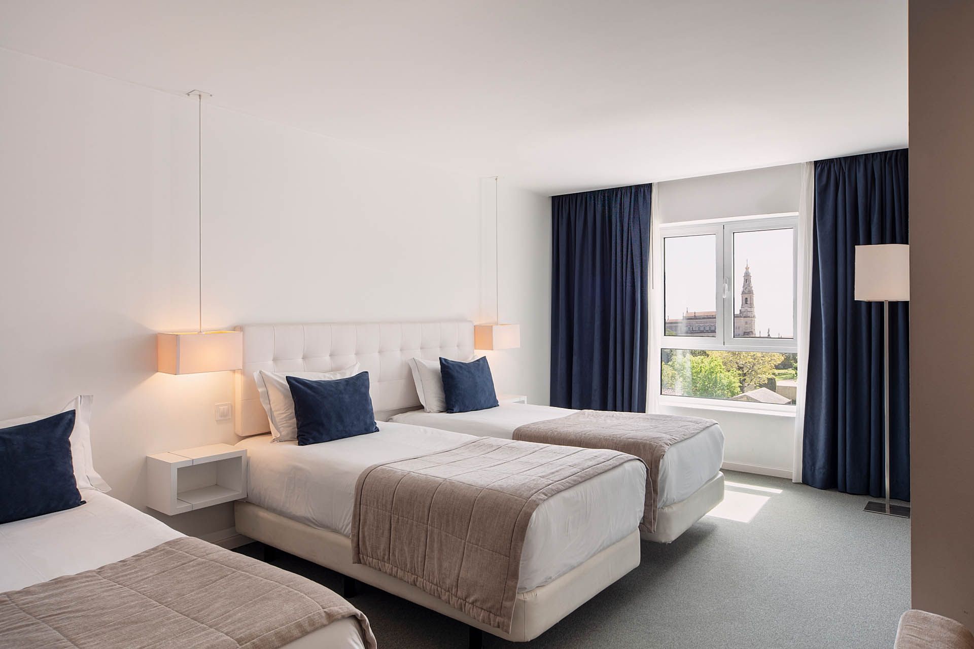 Quarto de hotel luminoso com três camas de solteiro, cortinas azuis e vista para a vegetação.