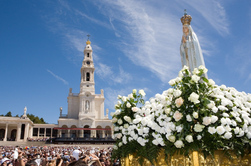 Praça lotada com a torre de uma igreja e uma estátua coberta de flores sob um céu azul.