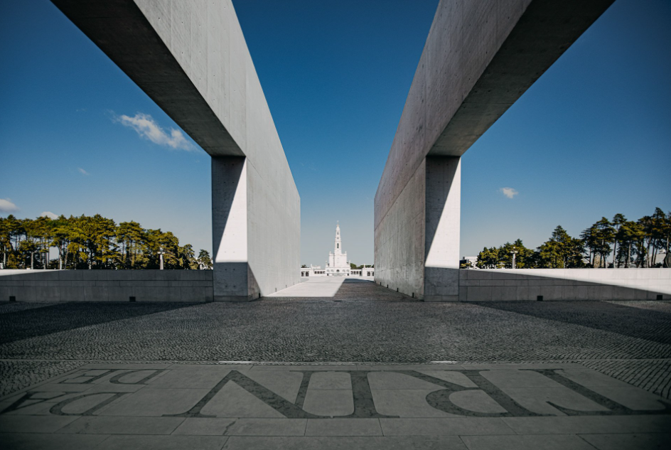 Entrada memorial moderna e simétrica com altas paredes de concreto e um monumento central sob um céu azul.
