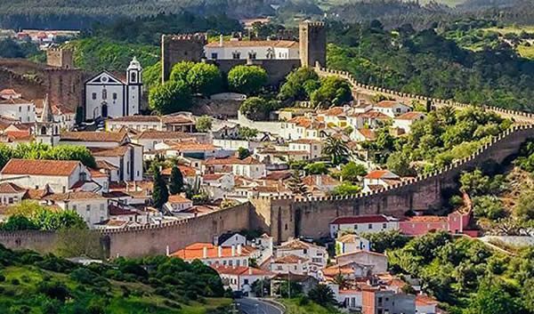 Cidade no topo de uma colina com edifícios de telhados vermelhos e uma grande muralha fortificada com vista para o vale.