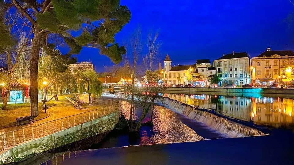 Cena noturna de um canal com prédios iluminados, árvores e reflexos coloridos na água.