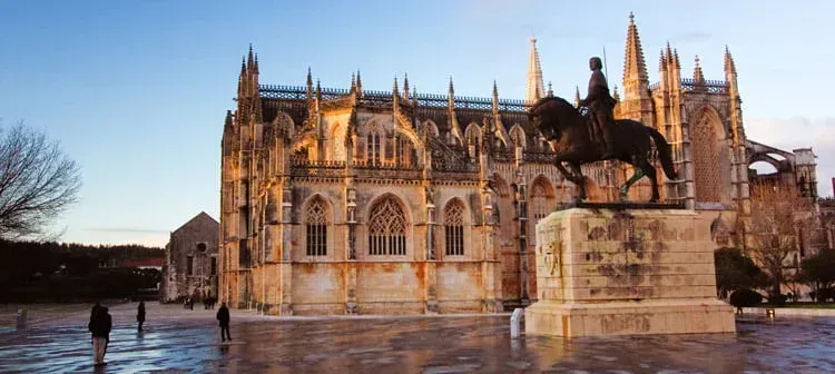 Catedral gótica com fachada de pedra ornamentada refletida em uma praça molhada ao pôr do sol, com algumas pessoas caminhando nas proximidades.